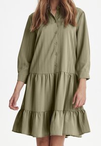 Zielono-oliwkowa sukienka z guzikami, z pionowymi paskami, długimi rękawami i rozkloszowanym falbaniastym dołem. Wykonana z gładkiego materiału o luźnym kroju.