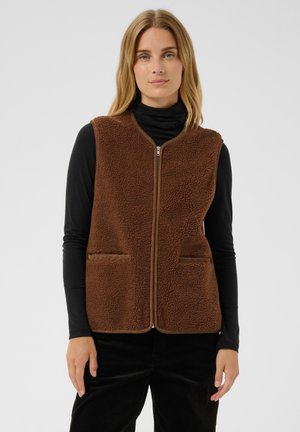 Femme portant une veste polaire marron zippée sur un haut noir à manches longues et un pantalon noir sur fond gris uni.