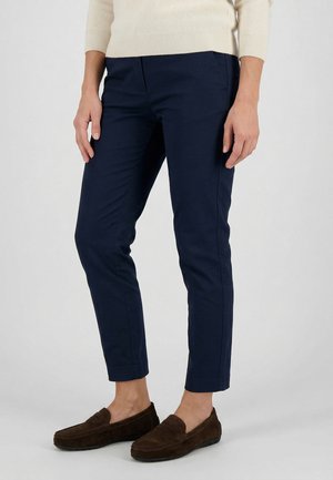 Kvinde iført navy slim-fit ankellange bukser med en beige sweater og brune loafers, stående mod en ensfarvet lys baggrund.