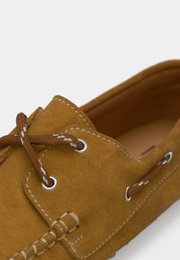 Zapato náutico de ante marrón con costuras blancas, ojales de metal y cordones marrones. El interior cuenta con un forro liso color marrón claro.