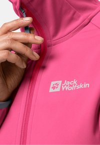 Ruka držící táhlo zipu jasně růžové bundy Jack Wolfskin s viditelným logem na hrudi.