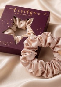 Chouchou en soie beige avec de petites perles décoratives, posé sur un tissu clair à côté d'un emballage violet foncé avec un texte doré et une fenêtre en forme d'étoile.