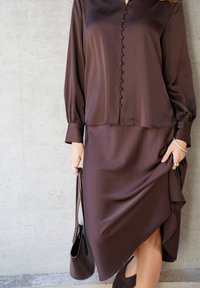 Ilga ruda satino suknelė su sagomis priekyje, pūstomis rankovėmis ir asimetrišku kraštu. Derinama su atitinkama ruda rankine ir sidabriniais žiedais.