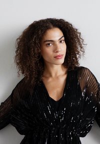 Blouse noire à sequins avec des manches transparentes, présentant des plis verticaux et un décolleté en V profond, exposée sur un fond clair.