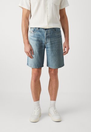 Mann trägt mittelblaue Jeansshorts, weiße Sneakers, weiße Sportsocken und ein kurzärmeliges weißes Hemd mit Knöpfen.