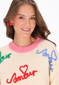 Donna sorridente con orecchini a forma di cuore rossi, che indossa un maglione crema con il testo colorato "Amour" e design a cuori, scollatura rosa e capelli castani sciolti.