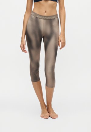 Femme portant un legging court marron brillant avec une ceinture "Peak Performance", debout pieds nus sur fond blanc.