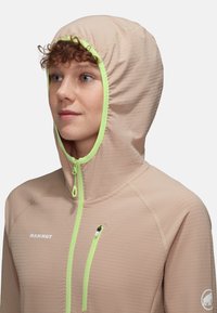 Leichte beigefarbene Jacke mit einer taillierten Kapuze, leuchtend grünen Akzenten, einer mit Reißverschluss versehenen Brusttasche und strukturiertem Stoff. Mit Logo auf der Brust.