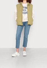 Persona vestida con chaleco sin mangas de color verde claro, sudadera color crema con estampado azul marino, jeans azules cortos y zapatillas blancas de pie sobre un suelo gris.