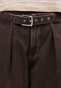 Κοντινό πλάνο σε σκούρο καφέ πλισέ παντελόνι με ταιριαστή ζώνη που έχει ασημένια αγκράφα και φαρδιά μπεζ μπλούζα μέσα από το παντελόνι.