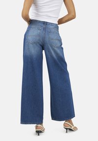Jeans in denim blu a gamba larga con lavaggio chiaro, dotati di due tasche posteriori, vita alta e orlo pulito. Indossati con tacchi aperti.