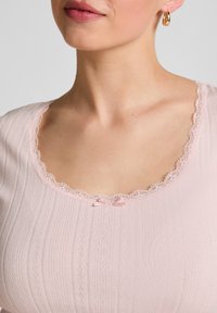 Lichtroze geribbelde top met een golvende kantenkraag en een klein strikdetail in het midden. Zachte stoftextuur, nauwsluitend ontwerp.