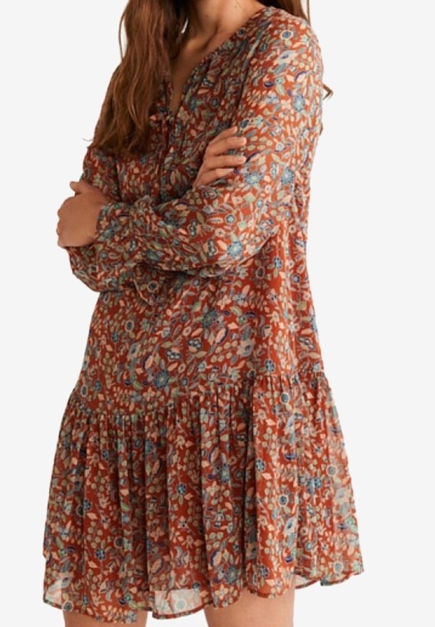 Femme portant une robe longue à manches longues couleur rouille à motifs floraux avec un ourlet à volants, debout, les bras croisés devant un fond uni.
