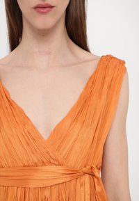 Orange klänning med omslag och djup V-ringning, tillverkad av texturerad tyg med veck, avslutad med ett knytbart midjebälte.