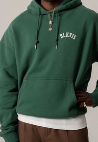 Hombre vestido con una sudadera verde de BLKVIS, camisa blanca debajo, pantalones marrones, anillo de oro y collares de oro en capas, frente a un fondo neutro.