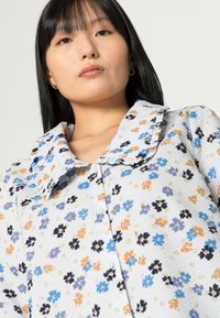 Camisa botão azul claro com um padrão floral em laranja, preto e azul. Apresenta um colarinho franzido e um tecido texturizado.