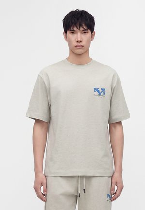Jeune homme portant un t-shirt beige clair et un pantalon assorti avec un logo bleu sur la poitrine et la cuisse, debout devant un fond blanc uni.