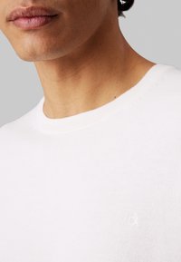Maglione bianco in cotone con collo a giro e texture a maglia fine, dettagli del logo ricamati sul petto. Tessuto liscio e design classico.
