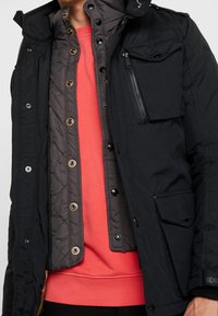 Svart ytterjacka med en zip-fick, tryckknappar och grått foder. Under syns en korallfärgad sweatshirt. Texturerat tyg genom hela plagget.