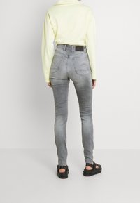 Jeans ajustados grises con un ligero desgaste, cintura alta y bolsillos traseros curvados. Combinados con una camisa amarilla de manga larga y sandalias negras.