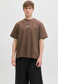 Brun bomulds T-shirt med korte ærmer, med rund hals og et trykt logo på brystet. Kombineret med løse sorte bukser.