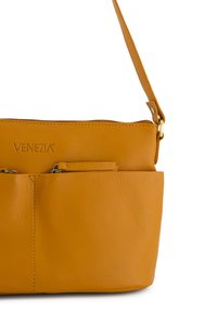Gul lædertaske med en glat overflade, der har en frontlomme og en lynlåslukning. Brandet med "VENEZIA" logoet på fronten.