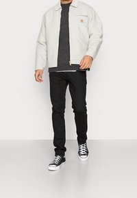 Giacca zip grigia chiara con colletto, maglione nero in maglia sotto, jeans neri e sneakers nere con dettagli bianchi.