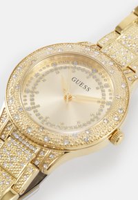 Goudkleurig horloge met een ronde wijzerplaat met kristallen accenten, een gestructureerde band en een gladde metallic afwerking. Merkennaam "Guess" prominent weergegeven.