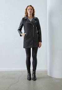 Cappotto grigio in misto lana con maniche in pelle nera, zip frontale e tasche laterali, abbinato a leggings in pelle nera e stivaletti con punta.