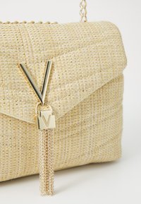 Svetąs, austǫ rašto rankinė su auksiniais akcentais, kuriame yra V formos metalinis detalis ir dekoratyvinis grandinėlės kutas. Tekstūruotas paviršius su subtiliu blizgesiu.