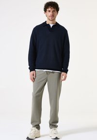 Marineblauer Strickpullover mit Kragen und V-Ausschnitt, kombiniert mit hellgrauen, schmal zulaufenden Hosen und beigen Sportschuhen. Einfarbiger Hintergrund.