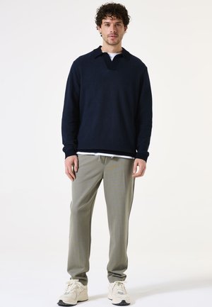 Marineblauwe gebreide pullover met een kraag en een V-hals ontwerp, gecombineerd met lichtgrijze tapered broeken en beige sportieve schoenen. Effen achtergrond.