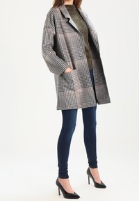 Manteau en plaid gris avec une coupe décontractée, de grandes poches et un col cranté, assorti à un jean bleu foncé et des talons noirs pointus.