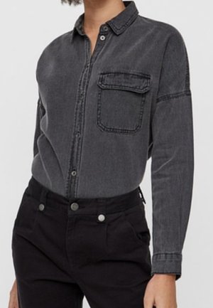 Kobieta ubrana w czarną, wypraną dżinsową koszulę z długim rękawem, z kieszenią z patką na przodzie oraz czarne, wysokim stanem spodnie.