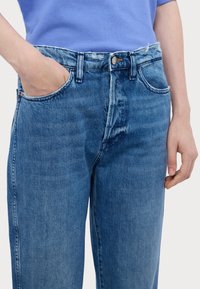 Vaaleansiniset farkut, joissa on korkea vyötärö, suora lahje ja näkyvät ompeleet. Ominaisuuksiin kuuluvat etutaskut ja metallinen nappikiinnitys.