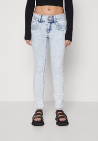 Žena oblečená v světle modrých skinny džínách s dvojitým zapínáním, černém dlouhém crop topu a černých sandálech na otevřenou špičku, stojí na bílé podlaze.