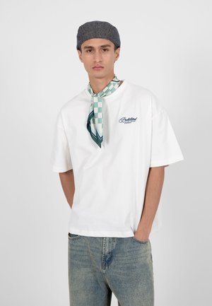 Ung mand iført grå kasket, hvid oversize T-shirt med logo, blå jeans og grønternet halstørklæde, stående med hænderne i lommerne.