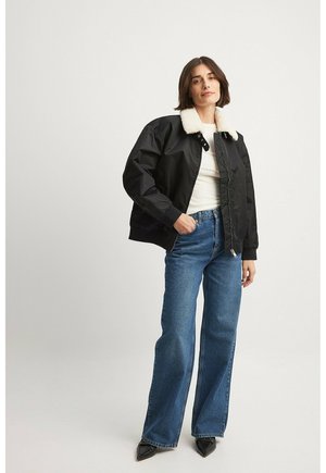 Chaqueta bomber negra con un cuello de piel sintética blanca, cierre de cremallera al frente, puños elásticos y un ajuste relajado, combinada con jeans acampanados azules y tacones negros.