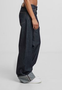 Tamsiai mėlynos denimo plačios kelnės su neapdirbtu kraštu, su baltomis siūlėmis ir odine etiketę gale. Dėvimos su baltais sportbačiais.