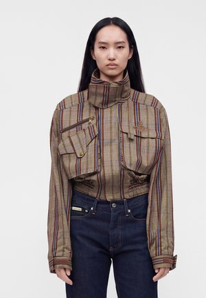Kvinde med langt mørkt hår iført en kort ternet jakke med høj krave og flere lommer samt mørkeblå jeans, der står foran en ensfarvet baggrund.