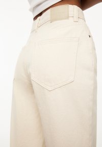 Pantalon en denim beige à jambes larges avec une finition texturée, doté de deux poches arrière et d'une étiquette en cuir de marque sur la ceinture.