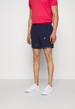 Marineblaue Shorts mit mehrfarbigen gestickten Polo-Logos, kombiniert mit einem roten Kurzarmhemd und weißen Sneakers, getragen von einem Model.