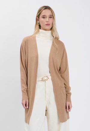 Vrouw draagt een beige open cardigan over een witte coltrui en hooggetailleerde broek met een witte riem, tegen een effen lichte achtergrond.