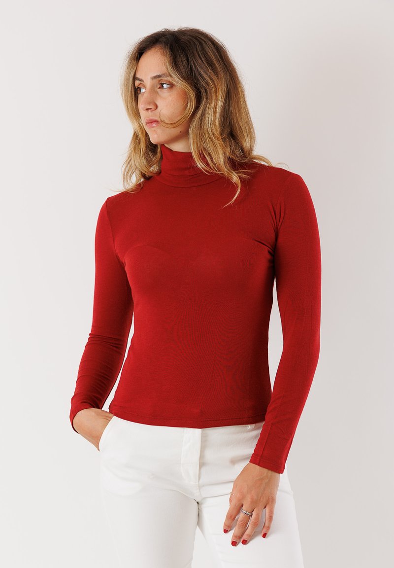 Maglietta rossa a maniche lunghe con collo alto in un morbido tessuto elasticizzato. Presenta un design aderente e una costruzione senza cuciture, abbinata a pantaloni bianchi.