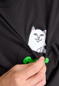 Black T-shirt featuring a cartoon white cat graphic with pink accents in a pocket. Hand pulling fabric reveals green smiley details beneath.