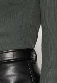 Grauer, gerippter Langarmoberteil mit gesticktem "G-Star Raw"-Logo und hochgeschnittenem, schwarzem Kunstledergürtel mit sichtbaren Nahtdetails.