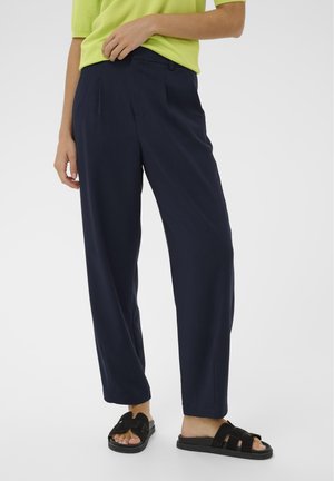 Pantaloni ampi color navy con pieghe frontali, realizzati in tessuto leggero. Abbinati a sandali neri con cinturini, indossati con una maglia verde brillante.