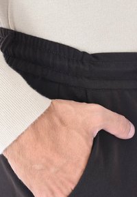 Pantaloni neri con un tessuto testurizzato, caratterizzati da una parte anteriore plissettata e un morbido elastico in vita. Una mano è posizionata nella tasca laterale.