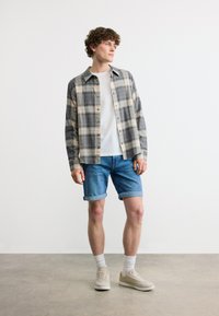 Νέος άνδρας που φοράει γκρι καρό πουκάμισο με κουμπιά πάνω από λευκό T-shirt, τζιν σορτς, λευκές κάλτσες και μπεζ αθλητικά παπούτσια, στέκεται σε τσιμεντένιο πάτωμα.