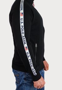 Felpa nera con zip e colletto alto, con il testo bianco "U.S. POLO ASSN." e la bandiera americana sulla striscia laterale. Texture del tessuto morbida.
